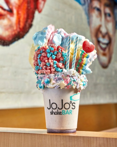 A colorful milkshake topped with cotton candy, candy pieces, and decorative straws, served in a JoJo's shakeBAR cup.
