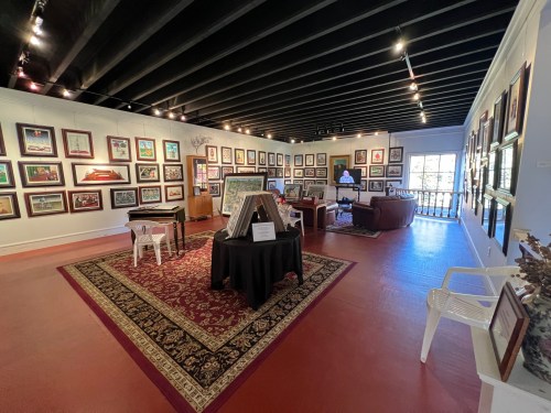 Art gallery interior featuring framed artworks on walls, a central display table, and seating areas.