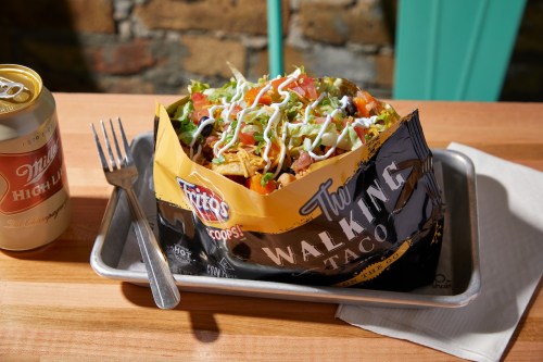 A "Walking Taco" in a Fritos bag topped with lettuce, tomatoes, and sour cream, alongside a can of beer on a wooden table.