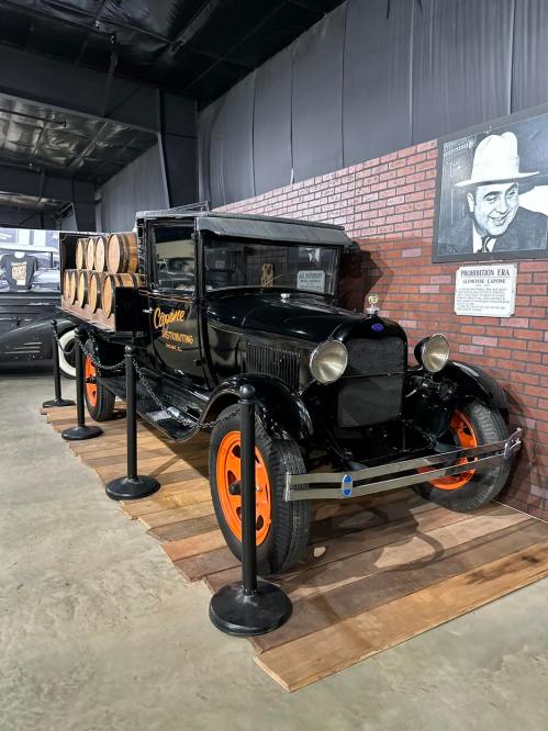 A vintage black truck with orange wheels, carrying barrels, displayed in a museum setting with a brick wall backdrop.