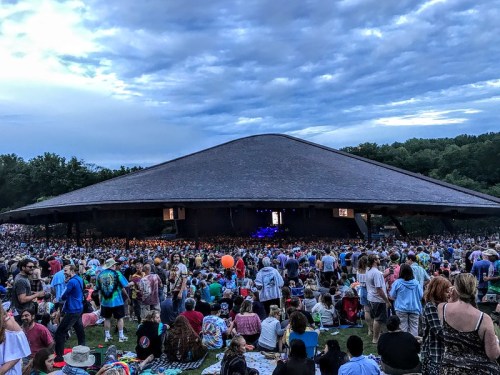 A large crowd gathers at an outdoor concert venue under a cloudy sky, with a stage visible in the background.
