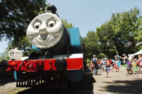 A smiling blue train with a friendly face, surrounded by children and families at a outdoor event.