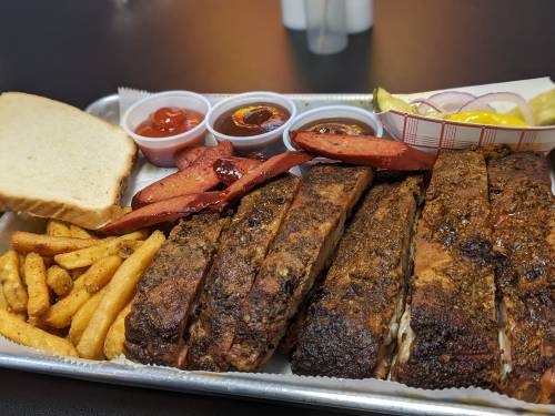 A platter of barbecue ribs, sausage, fries, bread, and various dipping sauces on a metal tray.