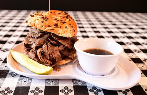 A stacked roast beef sandwich on a bun, served with a side of dipping sauce and a pickle slice on a checkered tablecloth.