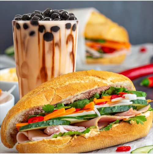 A close-up of a fresh sandwich filled with vegetables and meats, alongside a glass of bubble tea.