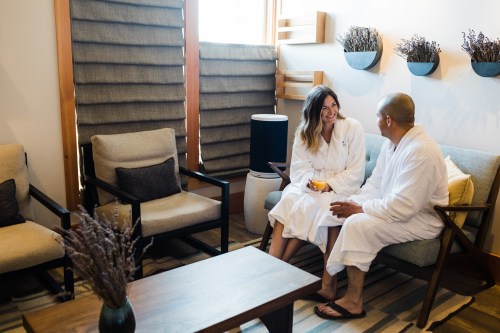 A couple in white robes sits in a cozy lounge, enjoying drinks and smiling at each other in a serene setting.