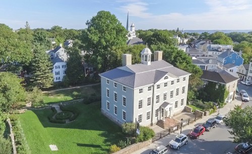 Aerial view of a historic building surrounded by greenery and residential homes, with a church steeple in the background.