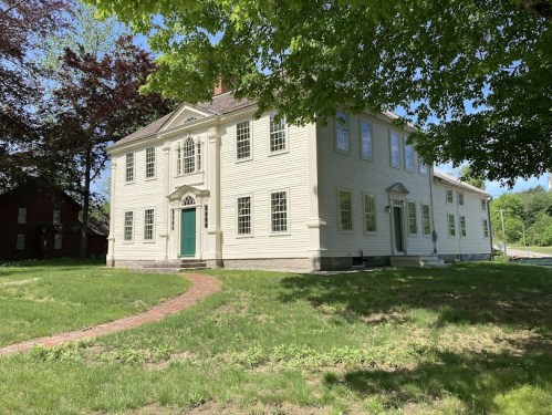 A large, historic two-story house with green doors and windows, surrounded by grass and trees on a sunny day.
