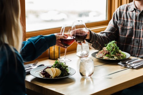 Two people clink wine glasses over a table with salads and water, enjoying a meal in a cozy restaurant setting.