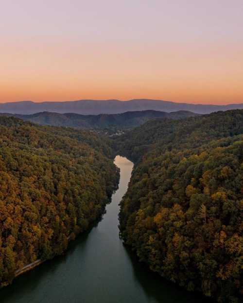 Aerial view of a winding river surrounded by lush, colorful forests at sunset, with mountains in the background.