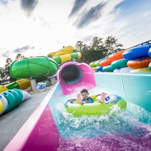 A child in a green inner tube slides down a water slide, splashing into a pool surrounded by colorful slides.