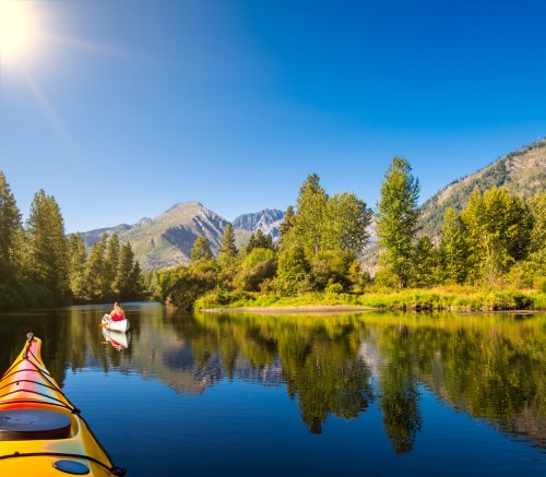 A serene lake scene with kayakers, surrounded by lush trees and mountains under a clear blue sky.