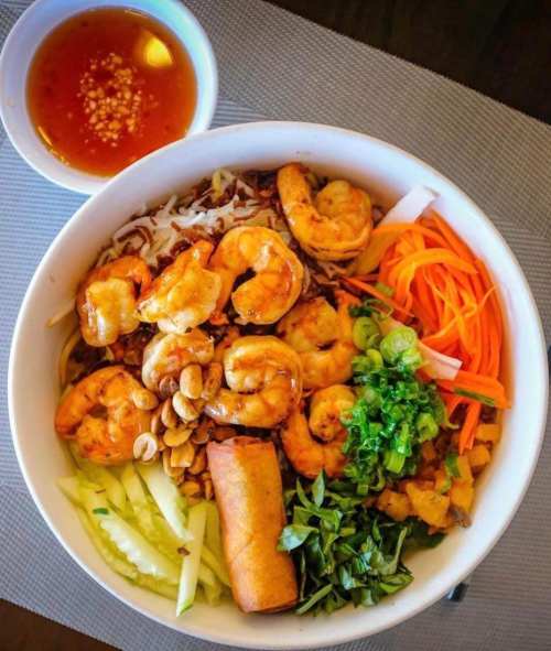 A bowl of shrimp vermicelli with vegetables, peanuts, and a spring roll, served with a side of dipping sauce.