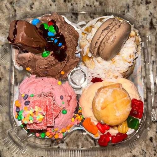 A clear container holds five colorful ice cream scoops topped with candies, macarons, and chocolate sauce.