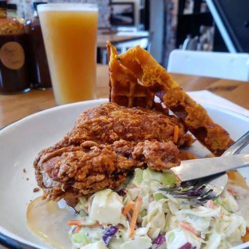 A plate with fried chicken, coleslaw, and a waffle, accompanied by a glass of orange beverage in the background.