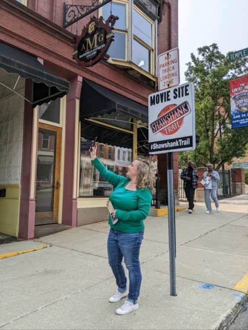 A woman in a green sweater takes a selfie in front of a building with a sign for the Shawshank Trail movie site.