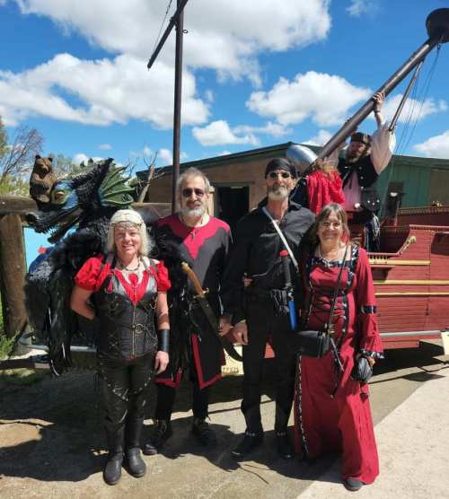 A group of four people in pirate costumes poses in front of a ship, with a dragon prop on one person's shoulder.