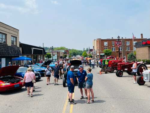 A bustling street filled with people admiring classic cars and tractors during a community event on a sunny day.