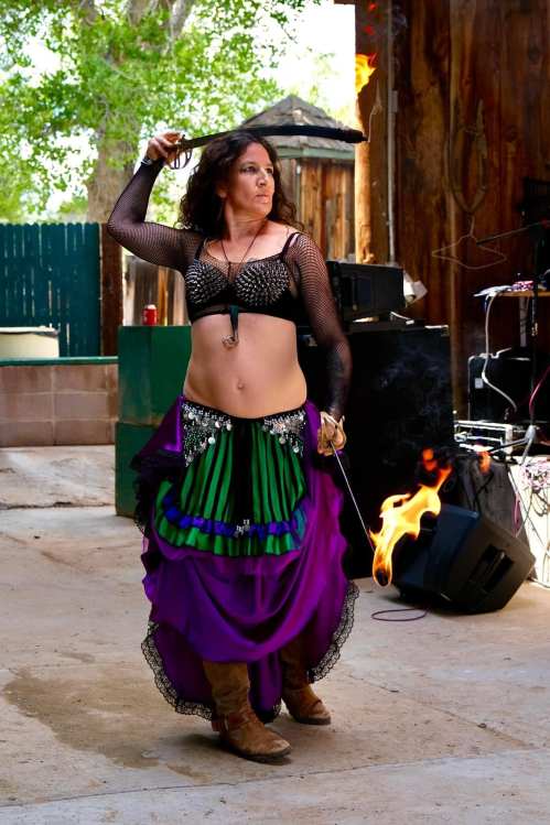 A dancer in a purple and green costume performs with a sword, surrounded by a lively outdoor setting.