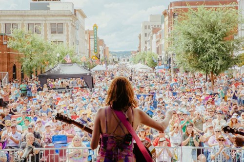 A musician performs on stage in front of a large crowd at an outdoor festival, with buildings and trees in the background.