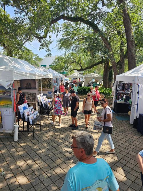 A bustling outdoor market with people browsing art and crafts under white tents, surrounded by trees.