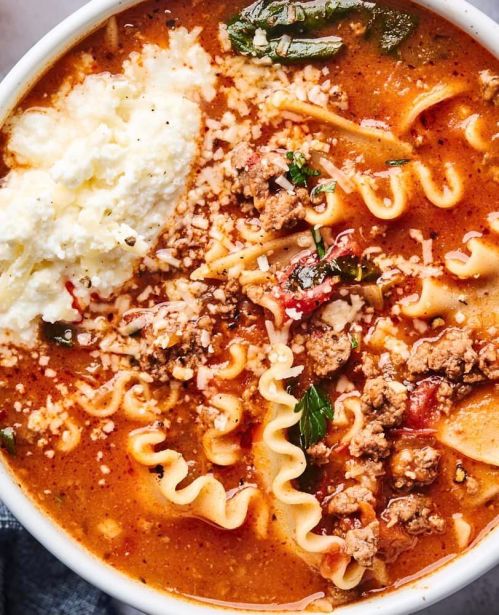 A bowl of hearty lasagna soup topped with ricotta, ground meat, herbs, and cheese, with wavy pasta noodles visible.