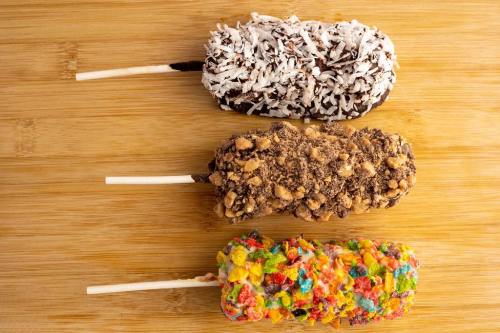 Three ice cream bars on sticks, coated in coconut, chocolate, and colorful cereal, arranged on a wooden surface.
