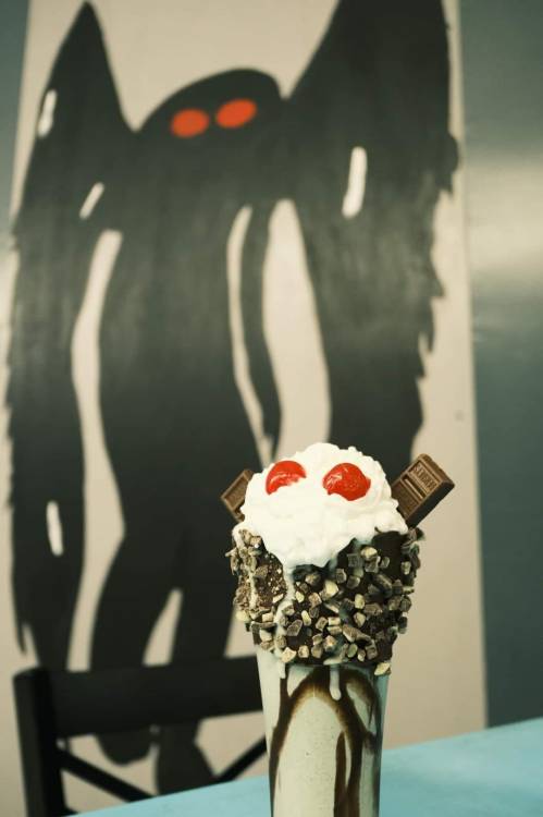 A decorated milkshake topped with whipped cream, cherries, and chocolate, with a shadowy figure in the background.