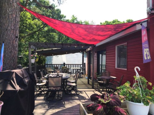 A patio with a red shade sail, surrounded by chairs and tables, next to a red building and greenery.