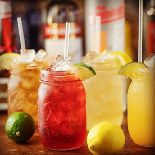 Four colorful cocktails in mason jars with ice, garnished with lemon and lime, on a wooden bar surface.