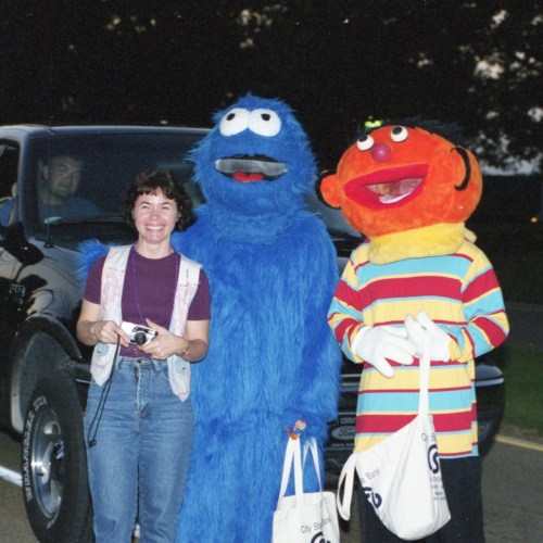 A woman poses with two costumed characters: a blue monster and a colorful, smiling character, near a vehicle.