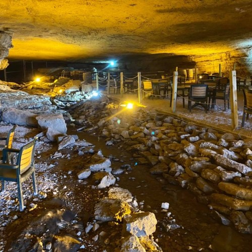 A dimly lit cave interior with a stream, stone pathway, and tables set up for dining.