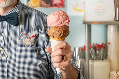 A person holding a cone with two scoops of ice cream: chocolate and strawberry, in a colorful ice cream shop.