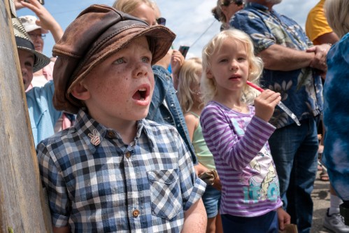 A young boy in a checkered shirt and hat looks surprised, while a girl in a striped shirt stands beside him, both watching intently.