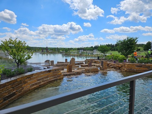 A serene landscape featuring a stone water feature, lush greenery, and a bright blue sky with fluffy clouds.