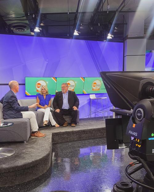 A television studio scene with three hosts seated on a couch, discussing in front of colorful screens.