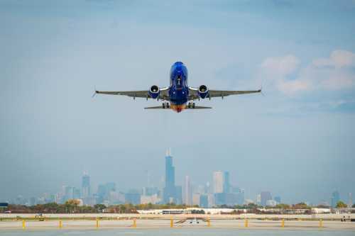 A plane takes off against a city skyline, with buildings visible in the background under a clear blue sky.