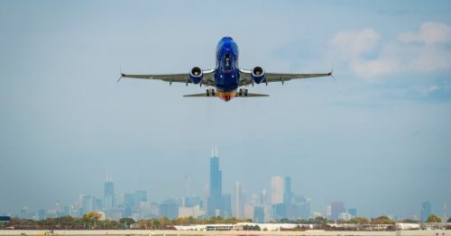 A plane takes off against a city skyline, with buildings visible in the background under a clear blue sky.