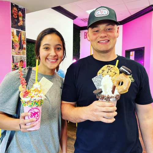 A young woman and man smile while holding colorful, oversized milkshakes topped with various treats in a vibrant shop.