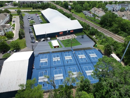 Aerial view of a tennis facility with multiple blue courts, surrounded by greenery and nearby buildings.
