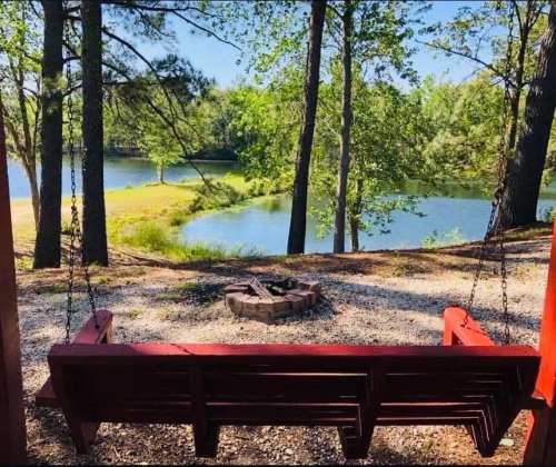 A red swing overlooks a serene lake surrounded by trees, with a fire pit nearby on a gravel path.