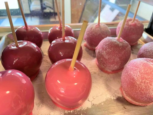 A tray of glossy red and pink candy apples, some coated in sugar, with wooden sticks inserted.