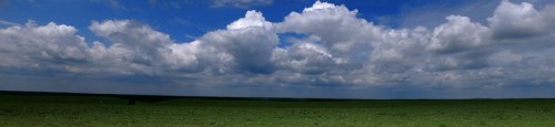 A panoramic view of a green landscape under a bright blue sky filled with fluffy white clouds.