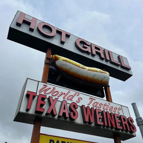 Sign for "Hot Grill" featuring a large hot dog and the text "World's Tastiest Texas Weiners." Cloudy sky in background.