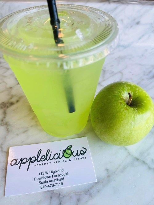 A green drink with a straw next to a green apple and a business card for "Applelicious Gourmet Apples & Treats."