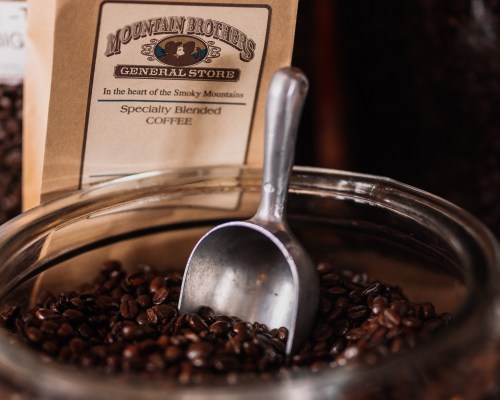 A close-up of coffee beans in a glass jar with a metal scoop, next to a bag labeled "Mountain Brothers General Store."
