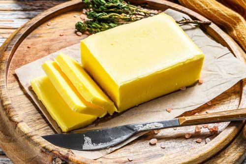 A block of butter with slices cut, placed on parchment paper on a wooden board, accompanied by herbs and a knife.