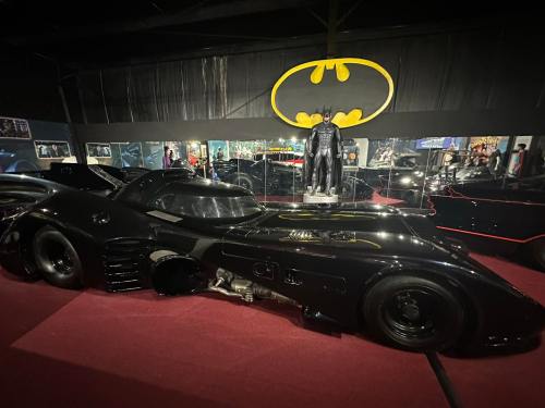 A sleek black Batmobile displayed in a dimly lit room with a Batman statue and iconic logo in the background.
