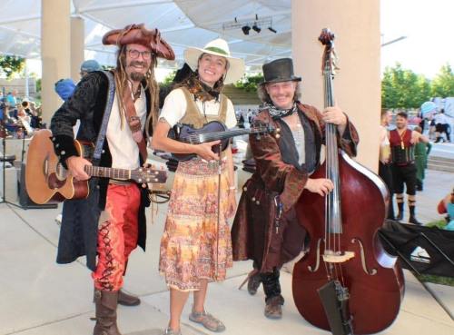 Three musicians in colorful costumes pose together outdoors, holding instruments and smiling at the camera.