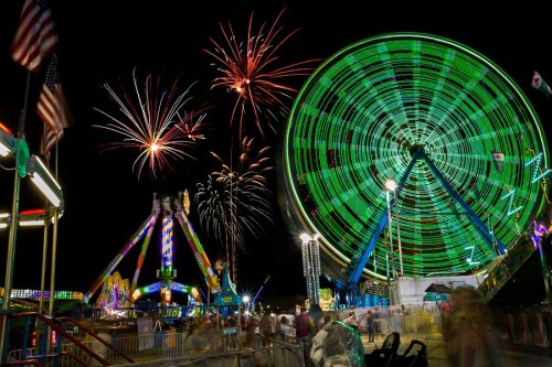 A vibrant carnival scene at night with a Ferris wheel, colorful lights, and fireworks in the sky.
